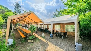 A covered outdoor dining area with tables and benches, surrounded by trees and a clear sky.