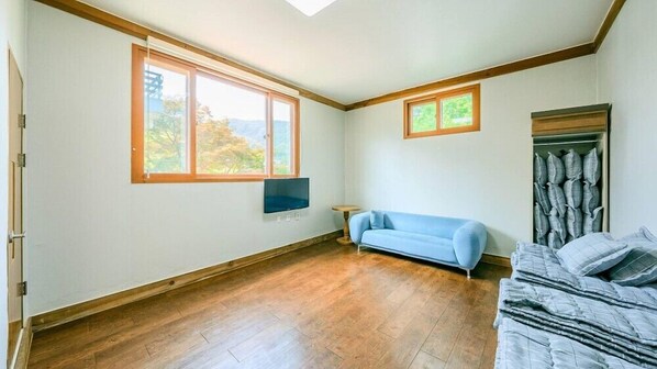 A room with a wooden floor, a blue sofa, a bed with a grey duvet, a TV mounted on the wall, and a window with a view of trees.