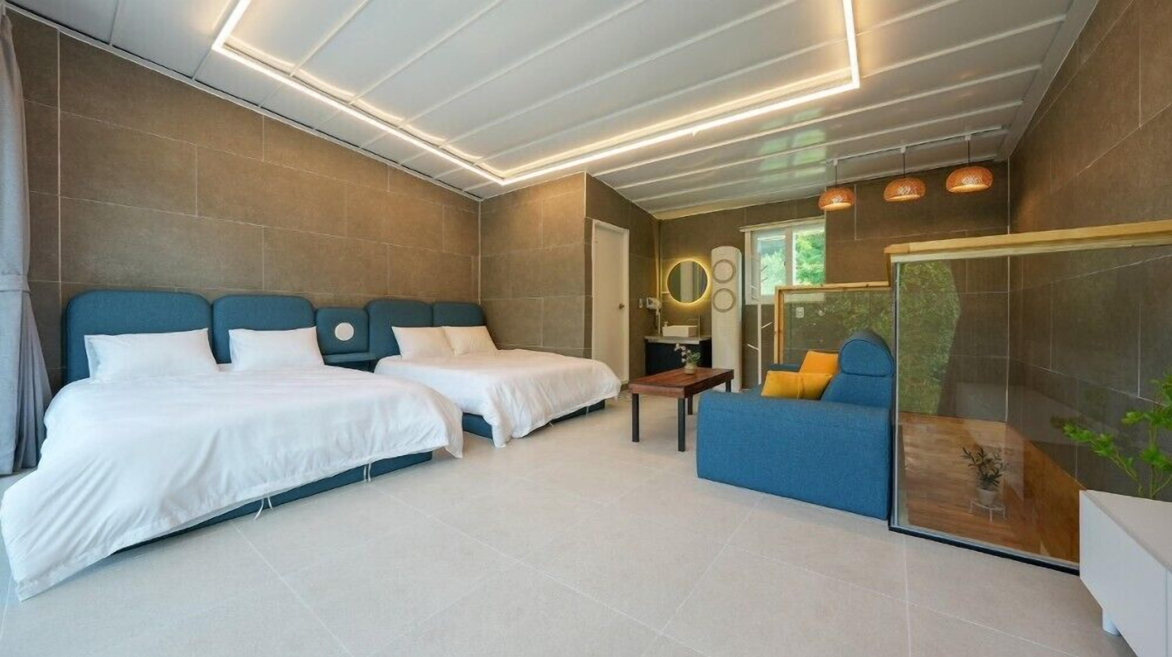 A modern hotel room with two beds, a sofa, a small table, and a glass partition.