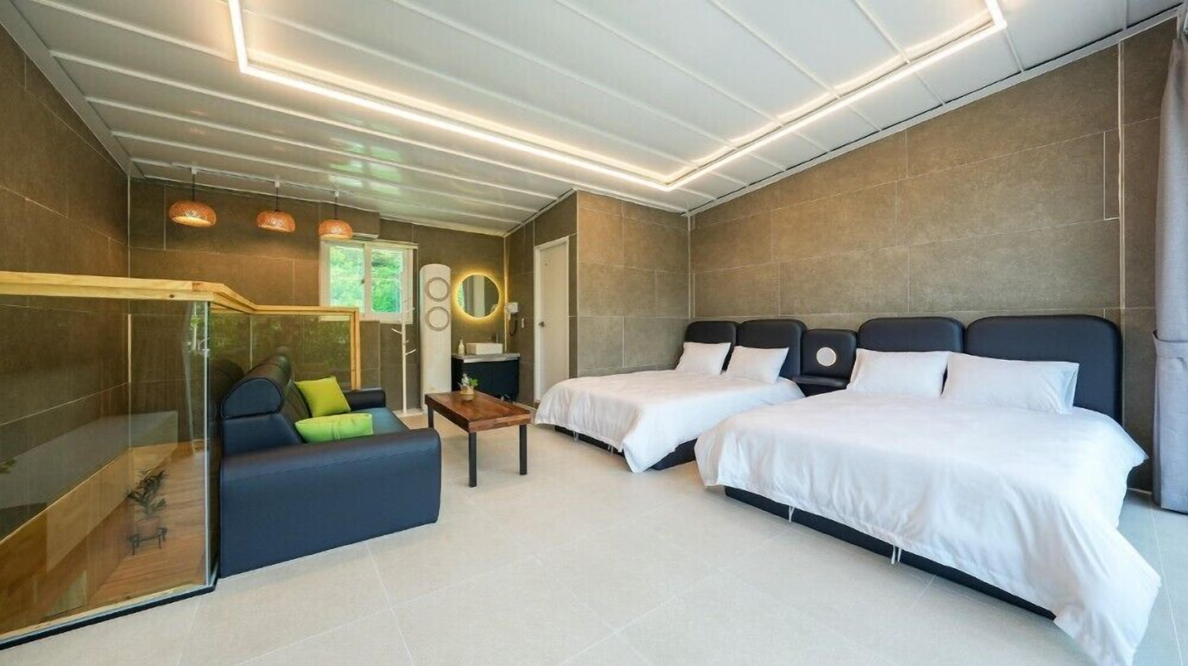 Una habitación de hotel moderna con dos camas, un sofá, una zona de ducha con paredes de vidrio y una mesita de noche con espejo y lámpara.