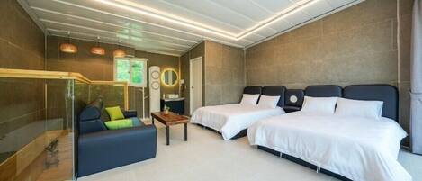 Una habitación de hotel moderna con dos camas, un sofá, una zona de ducha con paredes de vidrio y una mesita de noche con espejo y lámpara.