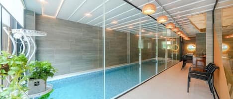 An indoor swimming pool with glass walls, modern lighting, and a seating area.