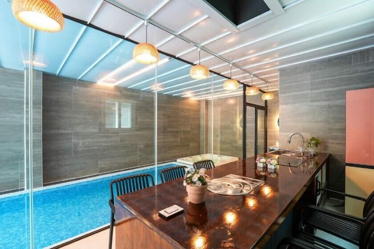 A modern kitchen with a large island, a built-in oven, and a sink. There is a glass partition separating the kitchen from a pool area.