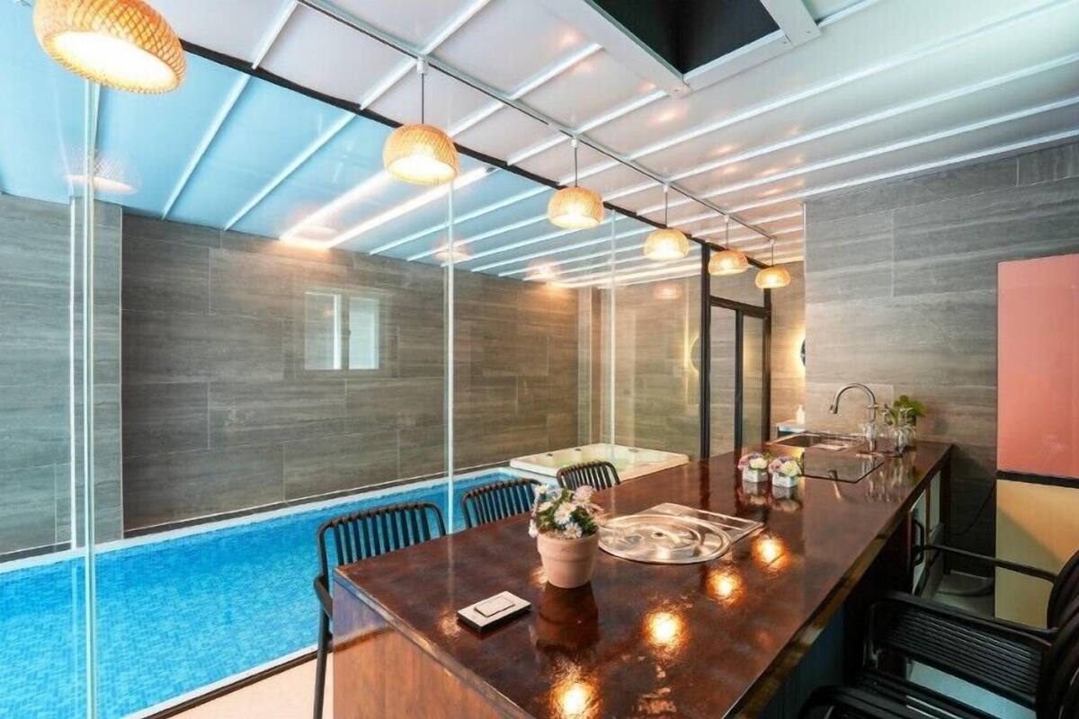 A modern kitchen with a large island, a built-in oven, and a sink. There is a glass partition separating the kitchen from a pool area.