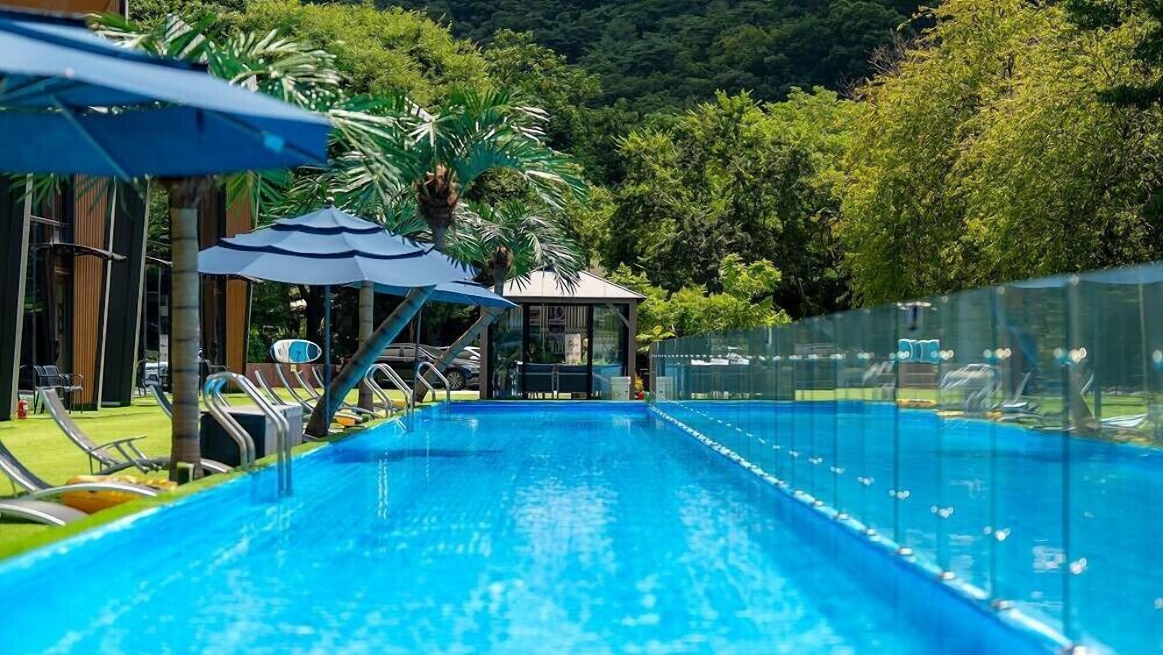 A swimming pool with lounge chairs and umbrellas, surrounded by a fence and trees.