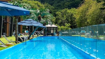 A swimming pool with lounge chairs and umbrellas, surrounded by a fence and trees.