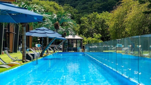 Une piscine entourée d’une clôture et d’arbres, avec des chaises longues et des parasols.