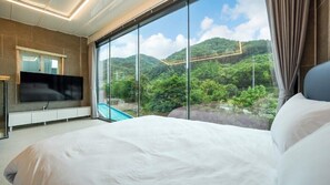 A bedroom with a large bed, a flat-screen TV, and a view of a lush green landscape through large windows.