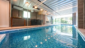 Piscina interior com água azul cristalina, área ensolarada com bar e assentos, e vista para o verde lá fora.