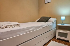 Ein Einzelbett mit weißer Matratze und einem gefalteten weißen Handtuch, ein Nachttisch mit Lampe sowie eine kleine, weiße Kommode.