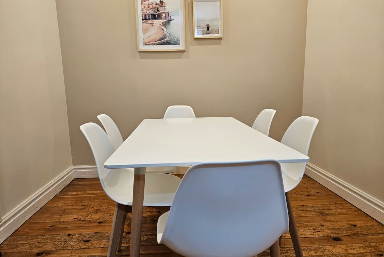 Una habitación con una mesa y sillas blancas, suelo de madera y dos cuadros en la pared.