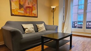 Un sofá gris con dos almohadas, una mesa negra, una lámpara de pie y un cuadro en la pared.