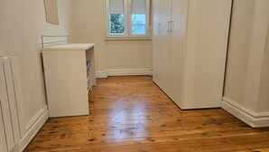 Una habitación estrecha con suelo de madera, paredes blancas, una ventana con persianas y un armario blanco.