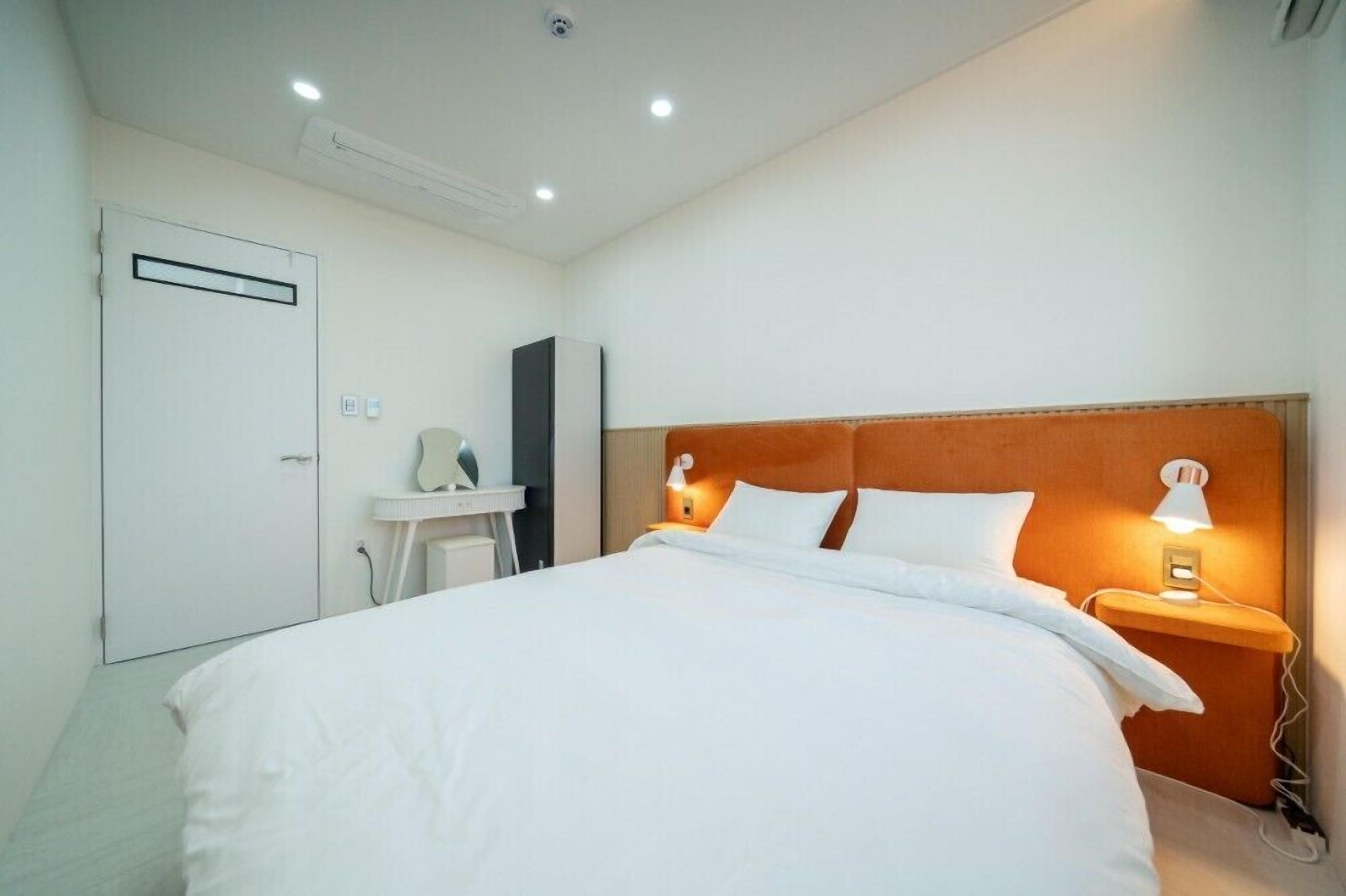 Habitación de hotel con una cama grande, una mesita de noche con lámpara, un armario y un pequeño escritorio con silla.