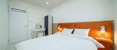 Habitación de hotel con una cama grande, una mesita de noche con lámpara, un armario y un pequeño escritorio con silla.