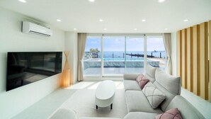 Una sala de estar moderna con un televisor de pantalla plana grande, una mesa de centro blanca y un sofá beige con cojines rosa. La habitación tiene ventanas amplias con vistas al océano y a una piscina.
