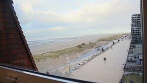 Une vue d’une plage avec un sentier de sable, quelques personnes qui se promènent et, au loin, un bâtiment au design moderne.