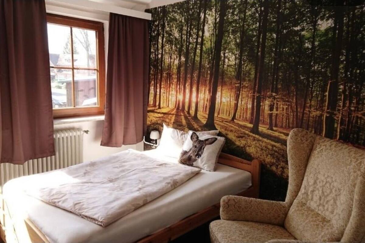 A bedroom with a bed, a chair, a window with curtains, and a forest-themed wall tapestry.