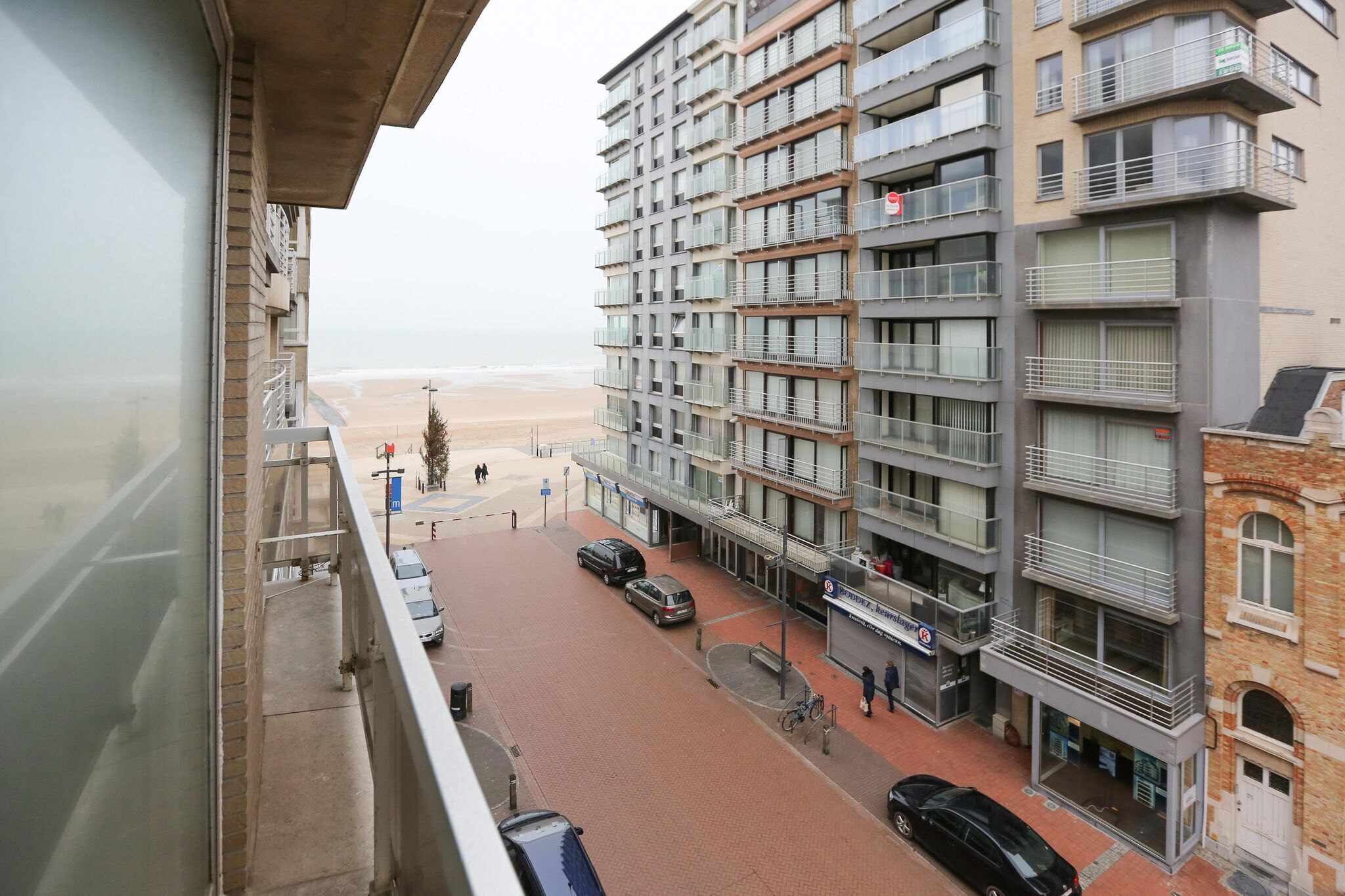 A multi-story building with balconies, a beach view, and a paved street with parked cars.