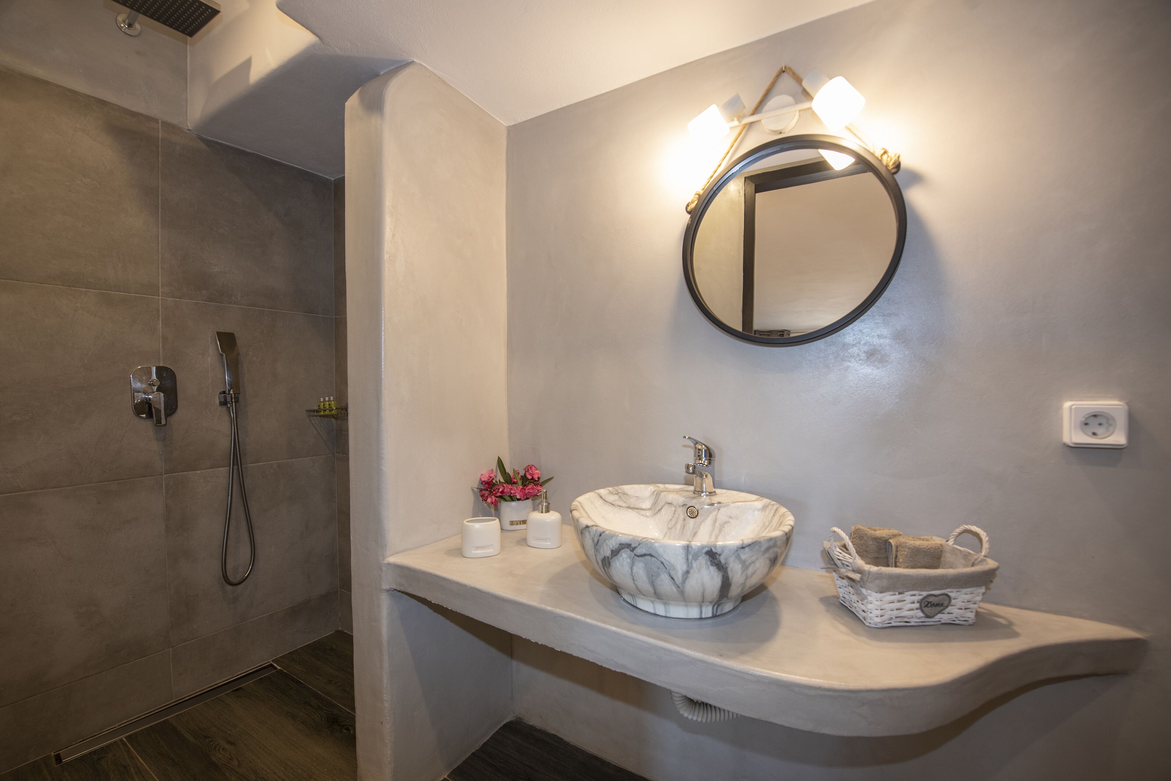 A bathroom with a marble sink, a round mirror, a handheld showerhead, and a small basket with towels.