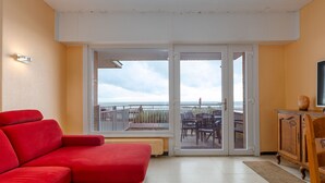 A room with a red sofa, a wooden cabinet, and a sliding glass door leading to a balcony with a view of the sea. - Ground floor apartment with sea view and garage (Middelkerke)