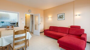 A living room with a red sofa, a dining table with chairs, a kitchen area with a microwave, and a door leading to another room. - Ground floor apartment with sea view and garage (Middelkerke)