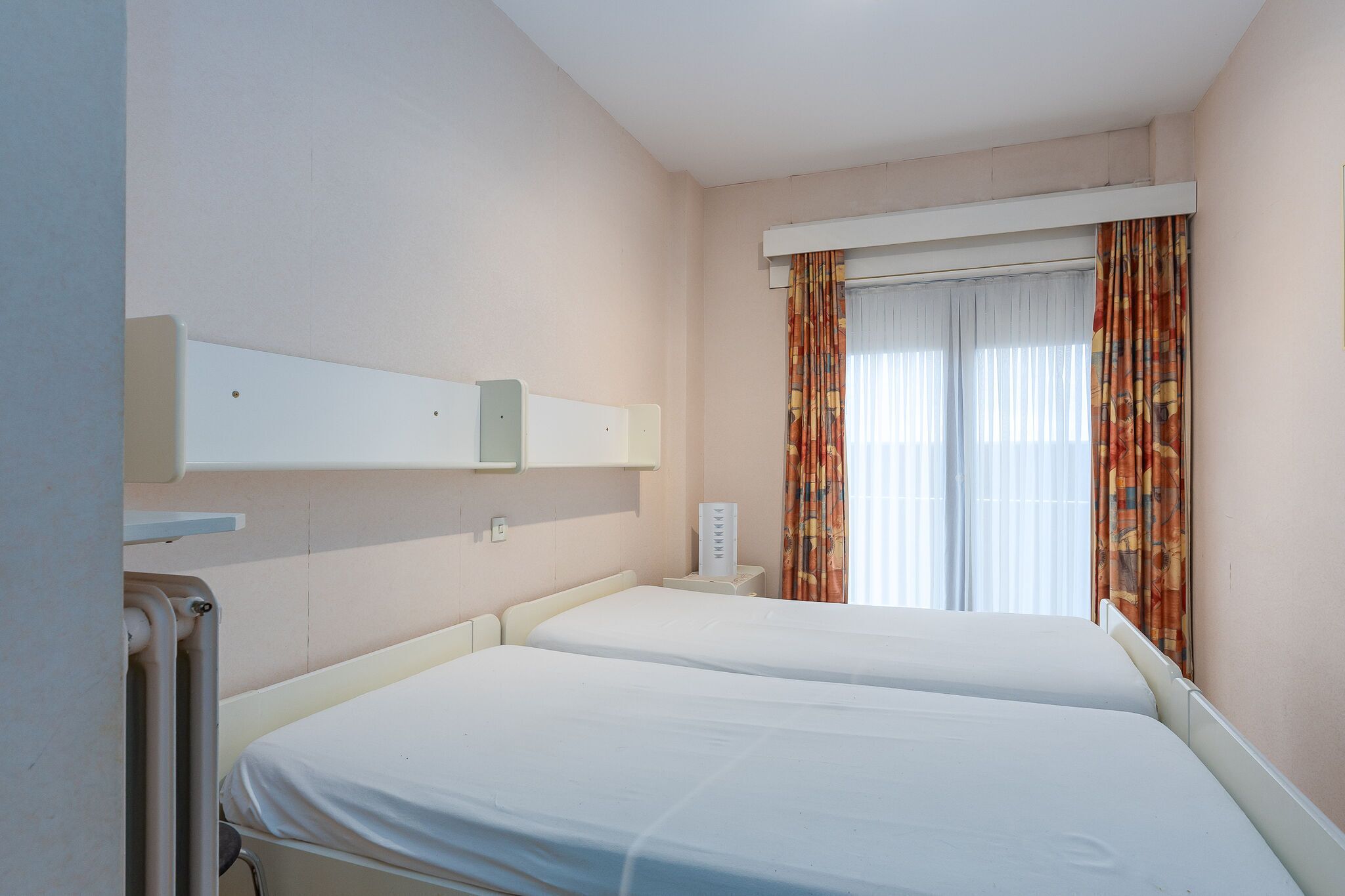 A hotel room with two beds, a window with curtains, a bedside table, and a wall-mounted shelf.