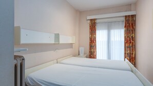 A hotel room with two beds, a window with curtains, a bedside table, and a wall-mounted shelf. - Ground floor apartment with sea view and garage (Middelkerke)