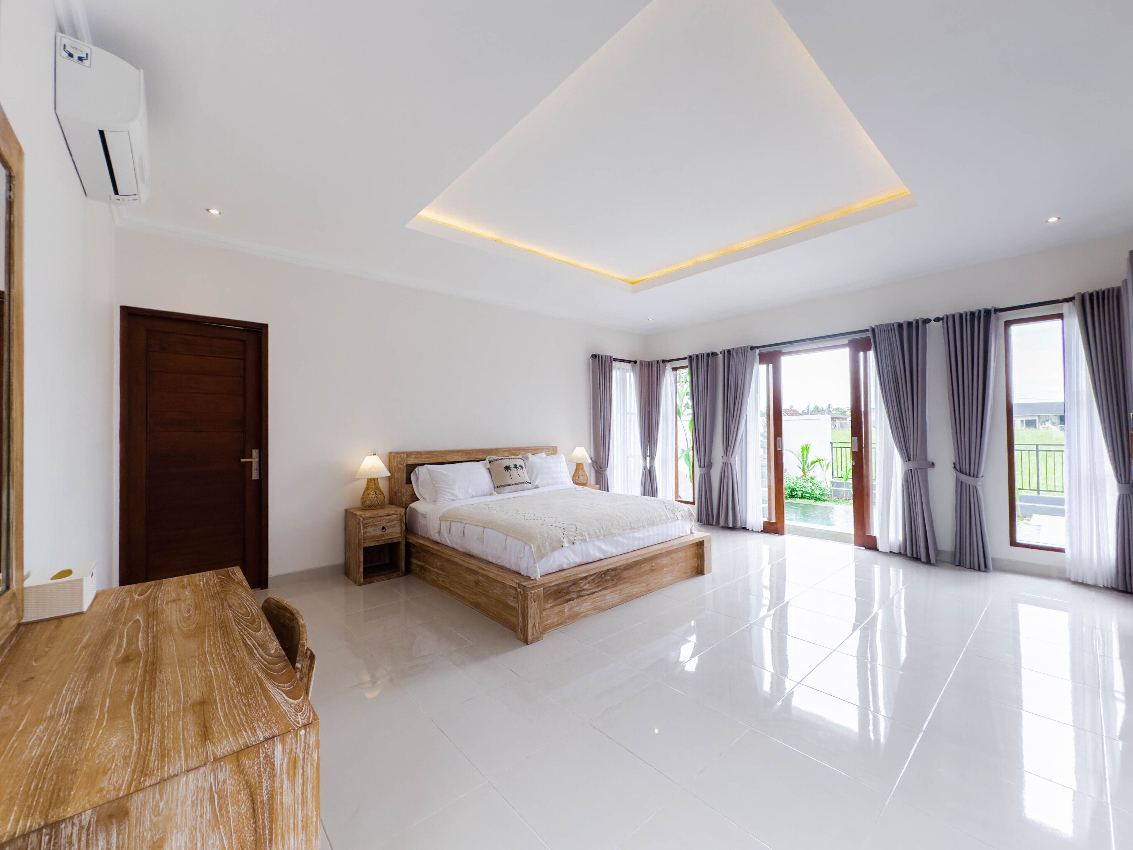 A spacious bedroom with a large bed, a wooden nightstand, a desk, and a view of a swimming pool.