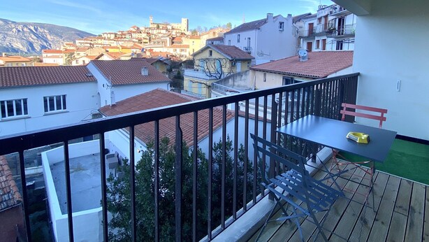A balcony with a table and a bench, overlooking a residential area with red-tiled roofs and a mountain in the background.
