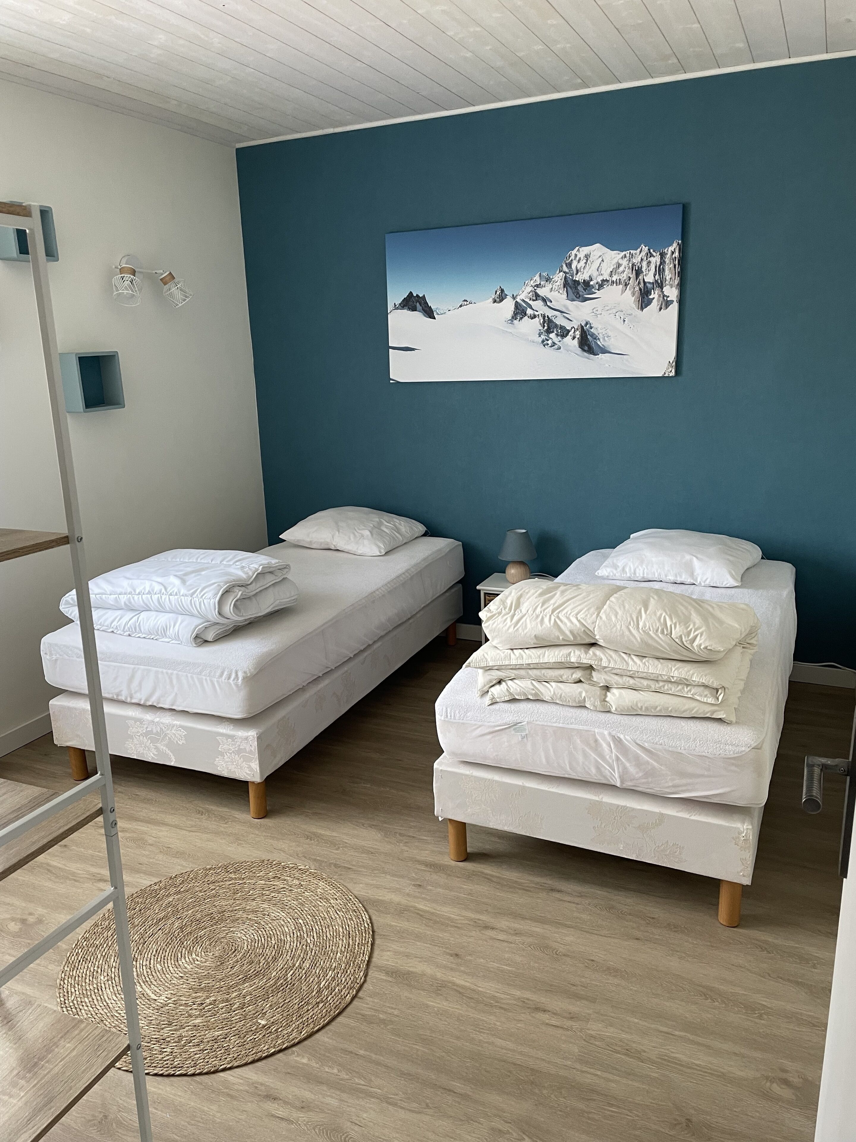 A bedroom with two beds, a blue accent wall, a mountain landscape painting, and a woven floor mat.