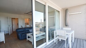 Una sala de estar moderna con un sofá de cuero azul, un ventanal que da a un balcón, y un juego de mesa y sillas blancos.
