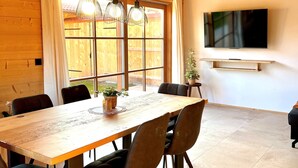 Una habitación con paneles de madera, una gran mesa de comedor, sillas, luces colgantes, un televisor y una ventana con cortinas.