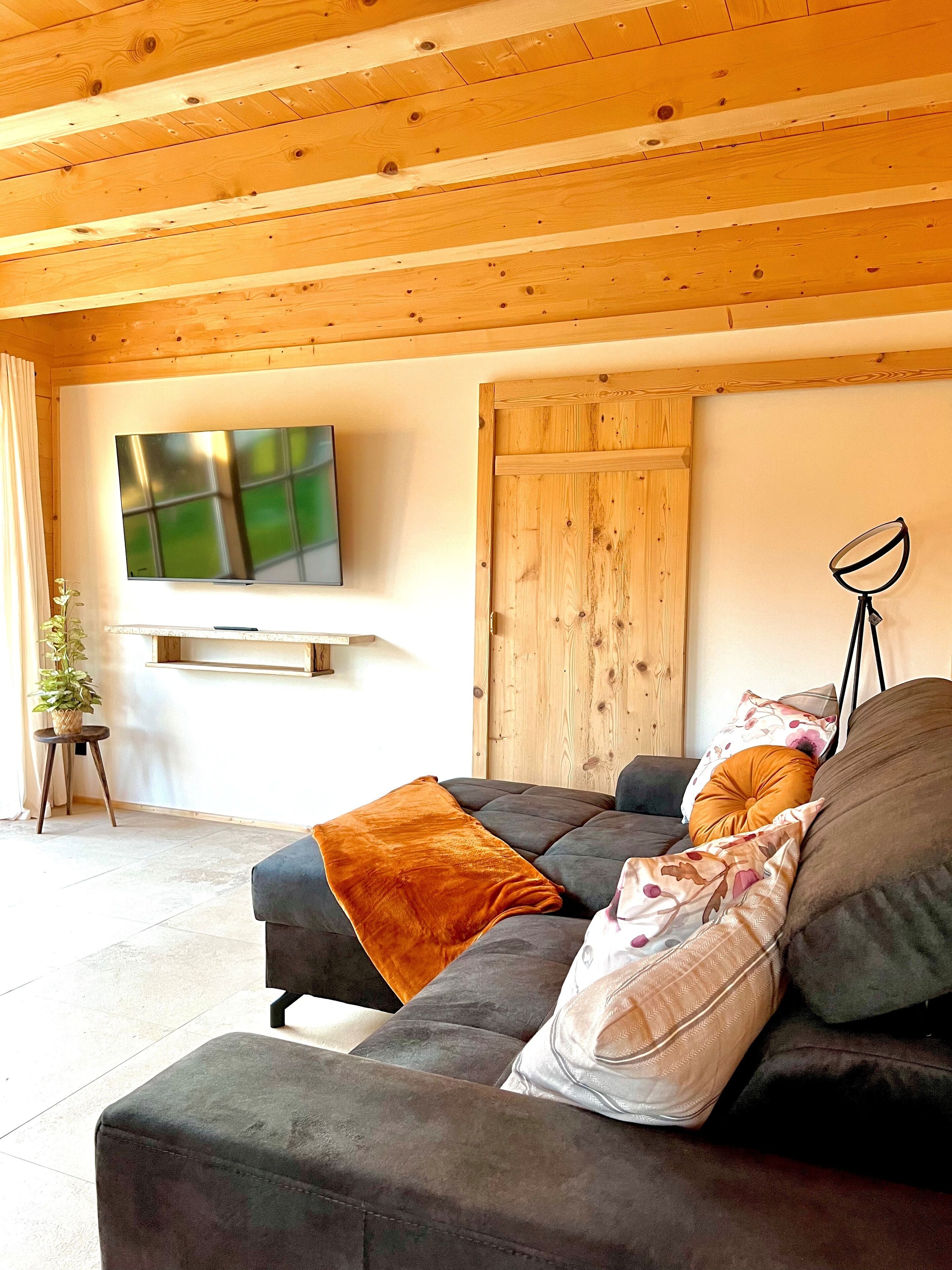 Un accogliente soggiorno con soffitto in legno, un'ampia TV a schermo piatto montata a parete, una porta in legno e una lampada da terra.