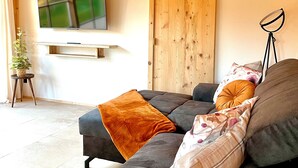 Una sala de estar acogedora con techo de madera, un televisor de pantalla plana grande montado en la pared, una puerta de madera y una lámpara de pie.