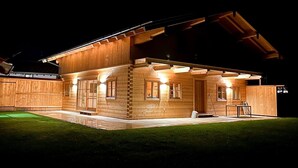 Una cabaña de madera con exterior iluminado, un porche cubierto y un pequeño espacio al aire libre con césped.