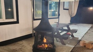 Un fogón de leña con recinto de vidrio, una mesa de picnic y una sombrilla en un patio al aire libre.