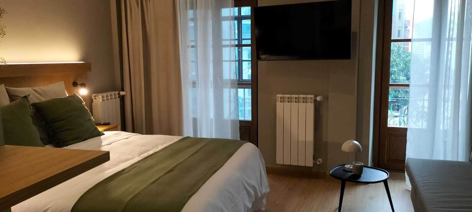 Une chambre d’hôtel comprenant un lit, une table de chevet, un téléviseur fixé au mur, un radiateur et une fenêtre avec des rideaux.