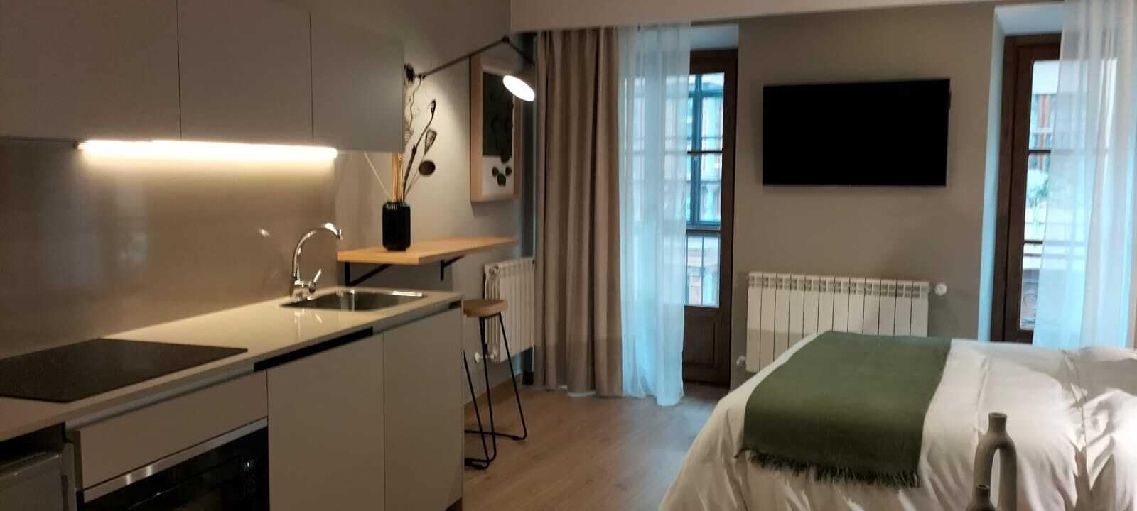 A modern kitchen with a built-in stove, a sink, and a small bar stool. There is a window with curtains, a radiator, and a flat-screen TV mounted on the wall.