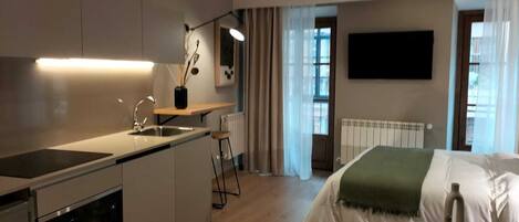 A modern kitchen with a built-in stove, a sink, and a small bar stool. There is a window with curtains, a radiator, and a flat-screen TV mounted on the wall.