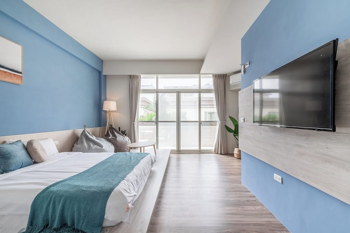 A modern hotel room with a large bed, a TV mounted on the wall, and a view of the outdoors through a large window.