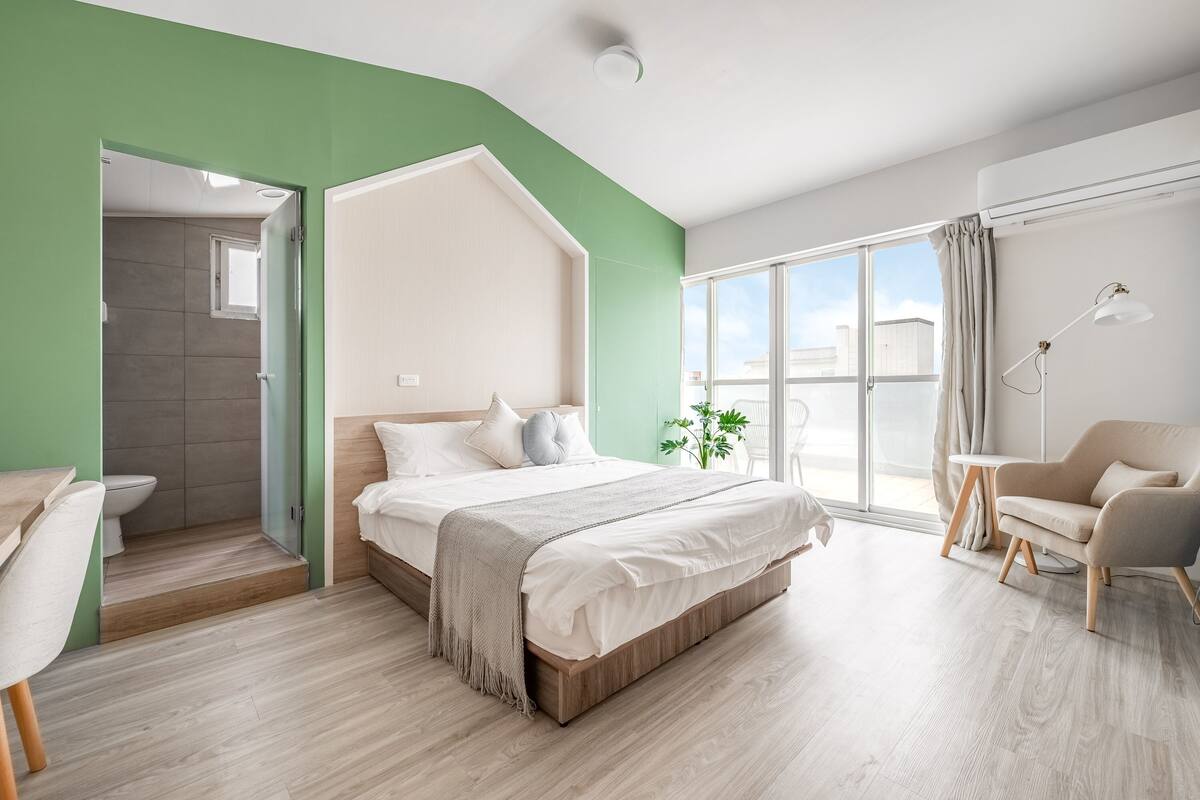 A modern hotel room with a large bed, a desk, a chair, and a view of the city through large windows.