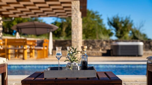 A wooden table with a tray containing a glass of water, a glass of wine, and a bottle of olive oil. In the background, there is a pool, outdoor seating, and a pergola. - Villa Oliveto with pool,jacuzzi & sauna (Donji Kašic)
