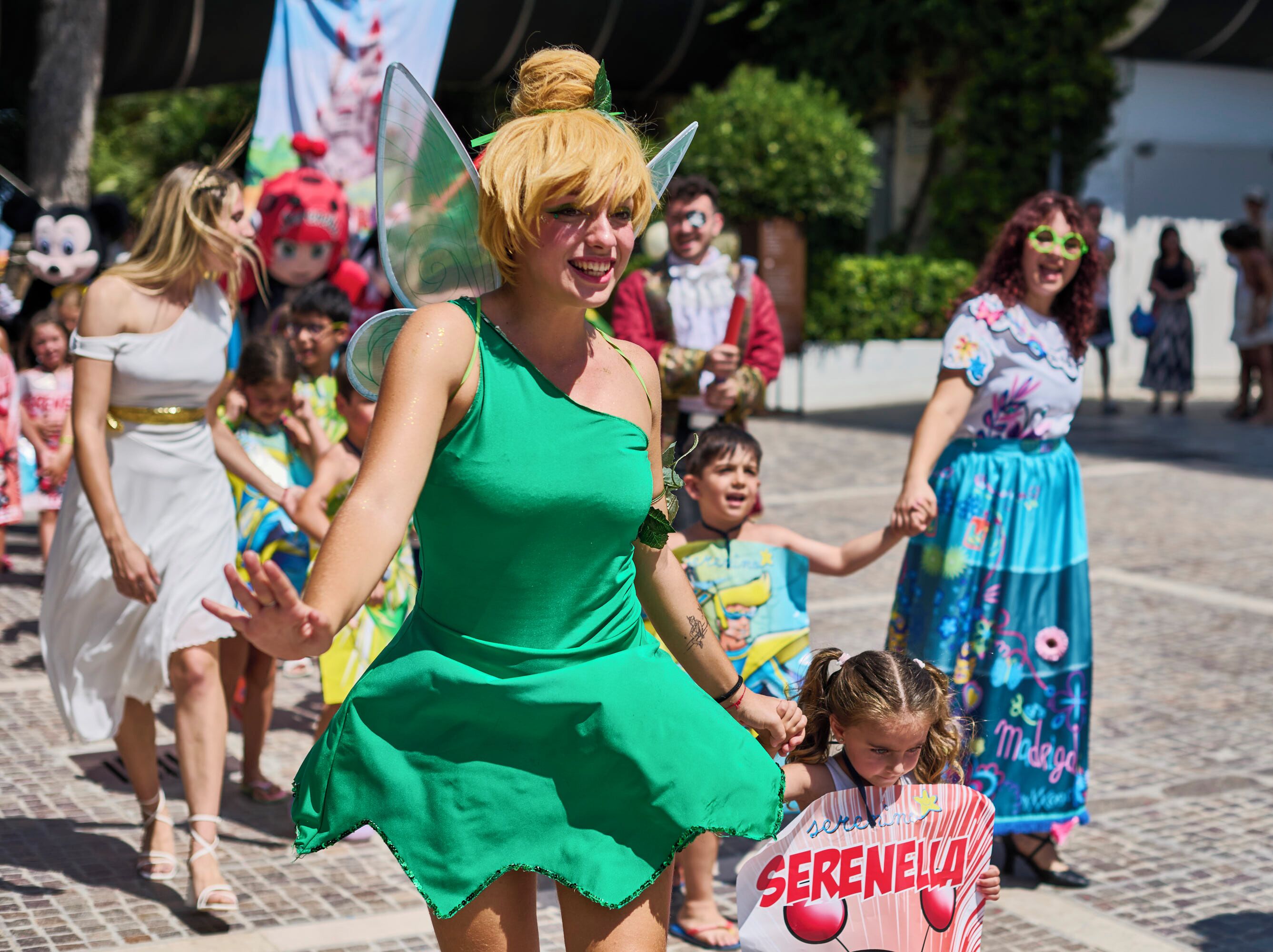 Una persona con un vestido verde y alas, un niño con una camiseta 'SERENELLA', y otro con una falda azul.