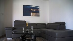 Een moderne woonkamer met een hoekbank, een glazen salontafel en een schilderij van een zonsondergang boven het water.