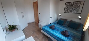 Une chambre à coucher comprenant un lit, une table de chevet, une commode, une plante en pot, une porte et une œuvre d’art accrochée au mur.
