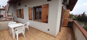 Un balcon avec une table et des chaises, des volets en bois aux fenêtres et une vue sur le quartier.