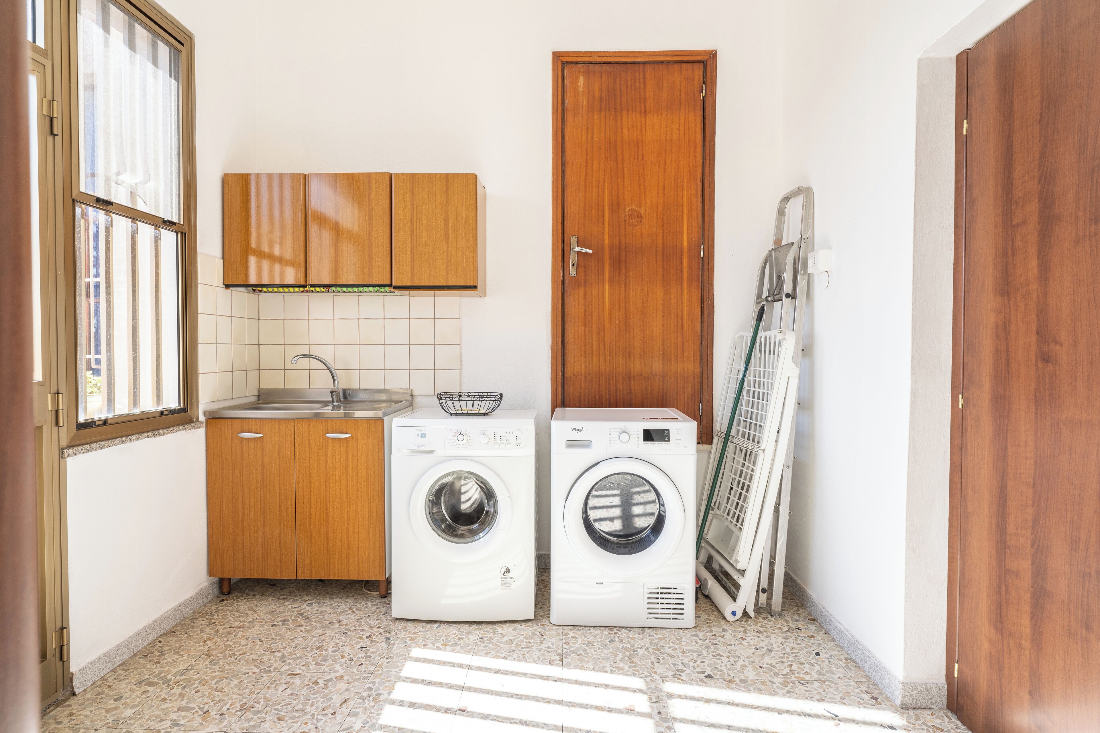 A compact laundry room with two washing machines, a wooden cabinet, a tiled backsplash, and a door.
