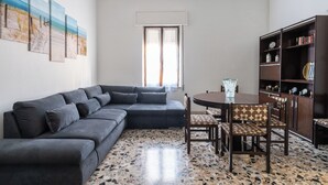 Una sala con un sofá seccional gris oscuro, una mesa redonda de comedor con sillas estampadas, una estantería y un ventanal con cortinas.