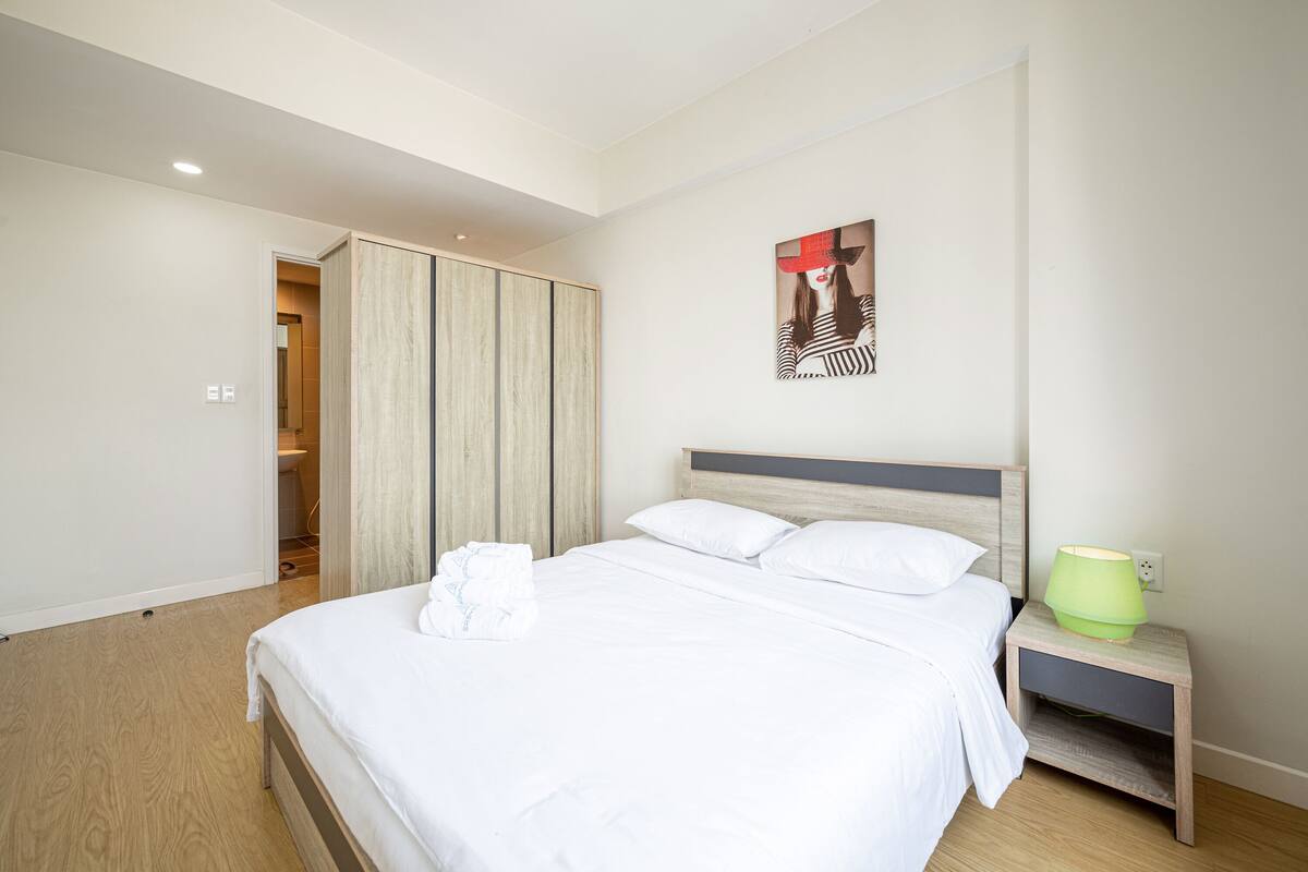 A modern bedroom with a large bed, a nightstand with a lamp, a wardrobe, and a painting on the wall.
